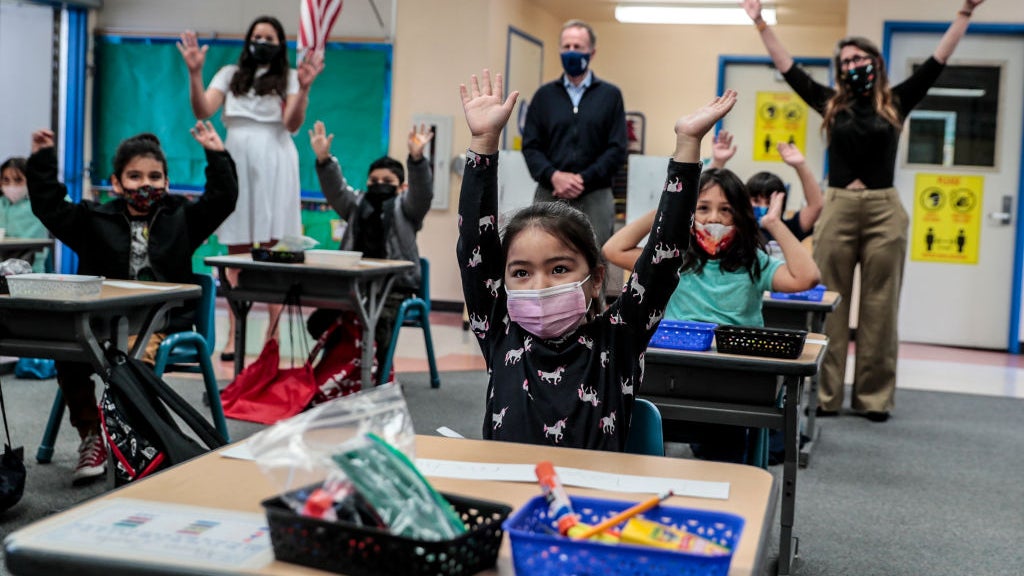 Group of excited masked kids in a kindergarten classroom with their hands raised above their heads