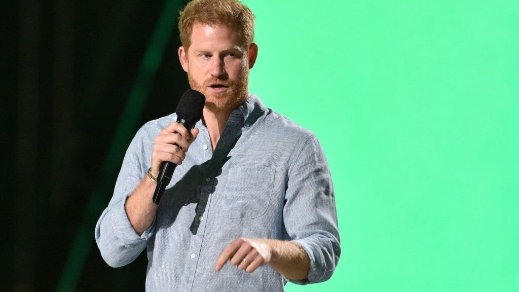 Prince Harry standing in front of black and green backdrop, holding a microphone and speaking.