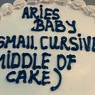 white cake with black icing reading "Aries Baby" and in parenthesis "small, cursive, middle of cake"