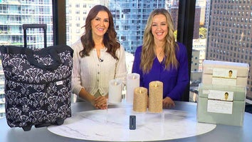 Two women standing behind a table displaying Carry-On Luggage, Luminara Flameless Candles, Kathy Ireland Sheet Set