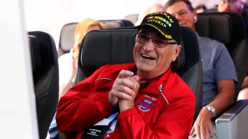 Elder man sitting in an airplane seat clapping and smiling wearing a red jacket and a Vietnam Veteran hat