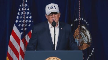 United States President Donald Trump speaking at a podium while wearing a navy blue suit and a white ball cap reading "USA"