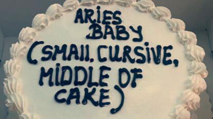 white cake with black icing reading "Aries Baby" and in parenthesis "small, cursive, middle of cake"