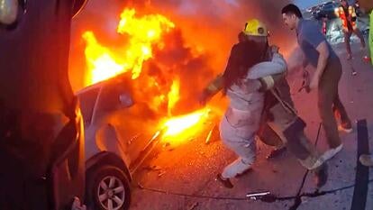 Bodycam image of a firey car with an officer pulling a woman from wreckage