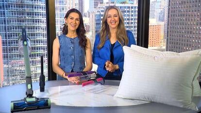 Two women behind a table displaying a vacuum, flat iron, and pillow
