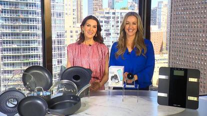 Two women standing behind a table displaying cookware, a charging dock, and a scale