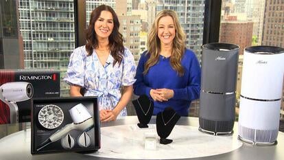 Two women standing behind a table displaying a hairdryer, necklaces, and air purifiers