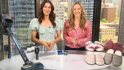 Two women standing behind a table displaying a vacuum, headphones, and slippers