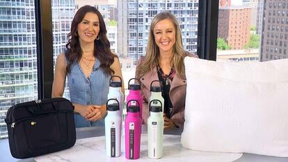 Two women standing behind a table displaying a tablet case, water bottles, and a pillow