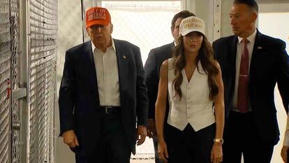 Donald Trump walking down an interior hallway along a chain link fence