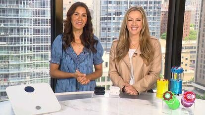 Two women standing behind a table displaying a smart scale, wireless earbuds, and lanterns