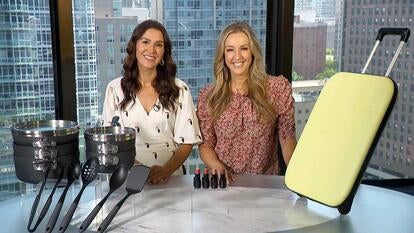 Two women standing behind a table displaying cookware, makeup, and a suitcase
