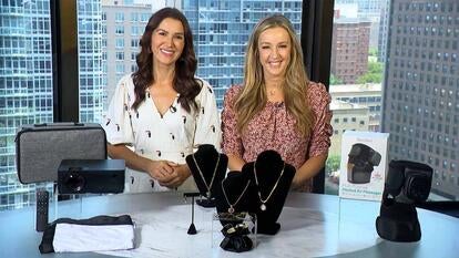 Two women standing behind a table displaying a projector, jewelry, and a massager