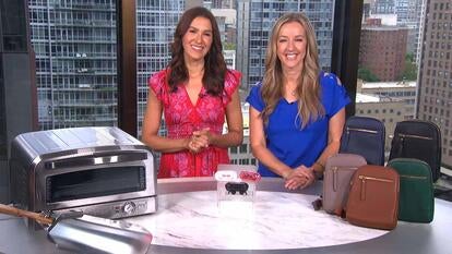 Two women standing behind a table displaying a pizza oven, earbuds, and a crossbody