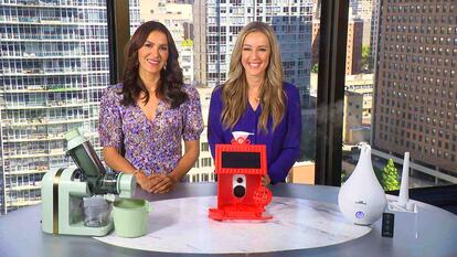 Two women standing behind a table displaying a Greater Goods Juicer, Hello Birdie Bird Feeder, Air Innovations Humidifier