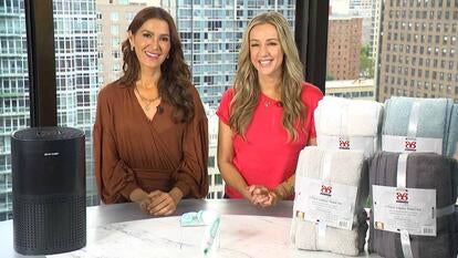 Two women standing behind a table displaying an Air Purifier, Facial Brush, Towel Set