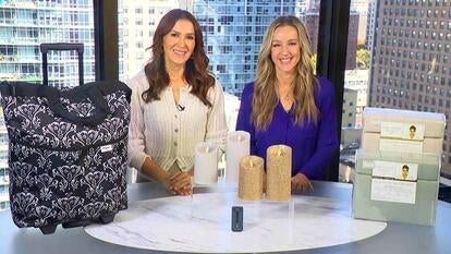 Two women standing behind a table displaying Carry-On Luggage, Luminara Flameless Candles, Kathy Ireland Sheet Set