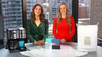 Two women standing behind a table displaying a Coffee Maker, Power Bank, Mattress Topper