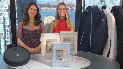 Two women standing behind a table displaying a Robot Vacuum, Sheet Set, Puffer Jacket