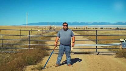 Man standing in front of a gate across a dirt road holding a sword 