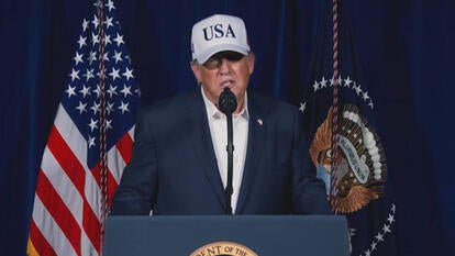 United States President Donald Trump speaking at a podium while wearing a navy blue suit and a white ball cap reading "USA"