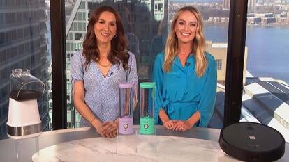 Two women standing behind a table displaying a Popcorn Maker, Portable Blender, Robot Vacuum