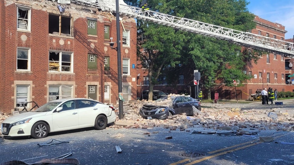 apartment building with broken windows, debris all over the street, top of building is destoyed, a car parked along the apartment building is destroyed by bricks that fell on top of it