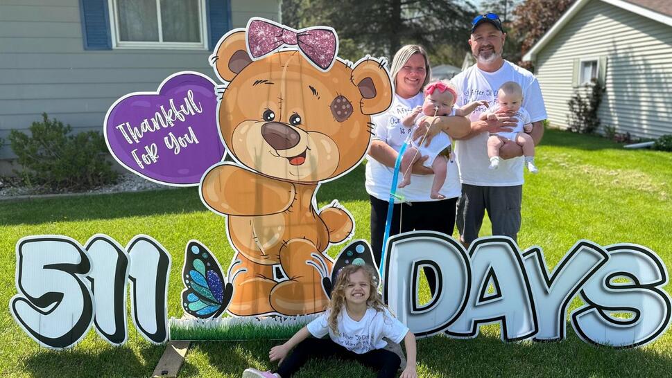 Katie Gatewood holding baby Charleigh and her husband Gary Gatewood holding their 11 month old baby and their oldest daughter on a lawn in front of a large sign os a teddy bear holding a heart that reads "Thankful for you" and text at the bottom reading "511 Days"