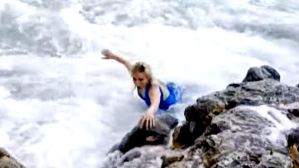 woman in blue dress holding onto rocks as waves hit her
