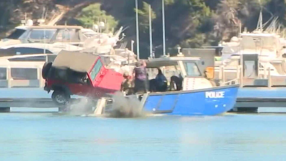 Red Jeep driving off of a jetty in front of a blue police boat