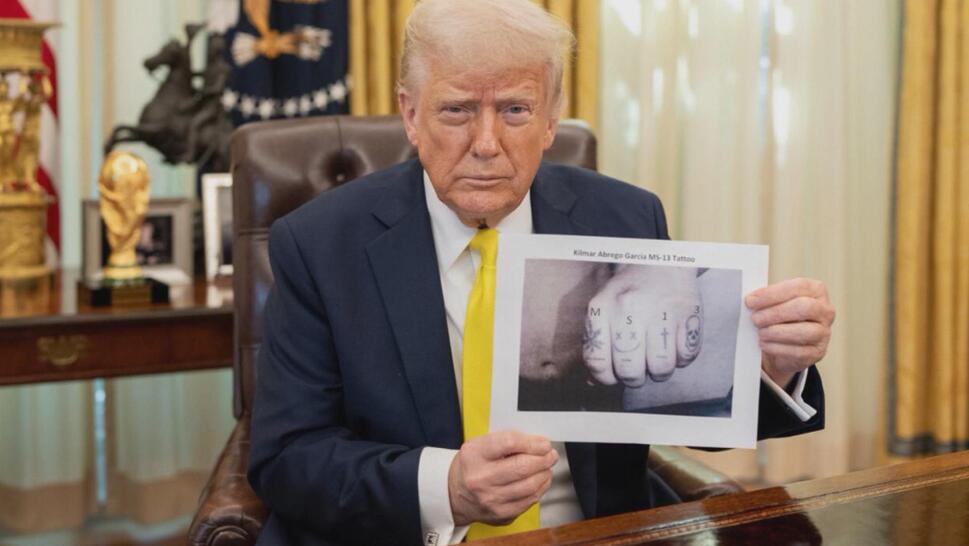 Donald Trump in the Oval Office, holding up picture of a tattooed hand with "MS13" photoshopped above the tattoos