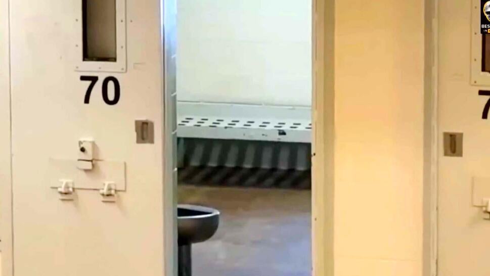 Open door of a prison cell showing the inside of a room with a bed and toilet