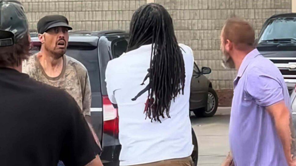A man in a white t-shirt and another in a purple t-shirt yell at a third man who is wearing a camo t-shirt and a baseball hat