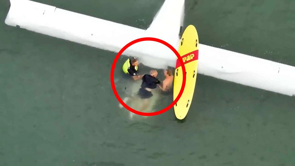 Birds eye view of a plane submerged in water as 2 rescuers hold a man above water