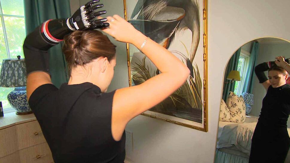 Teen with prosthetic arm looking into a mirror as she ties up her hair.