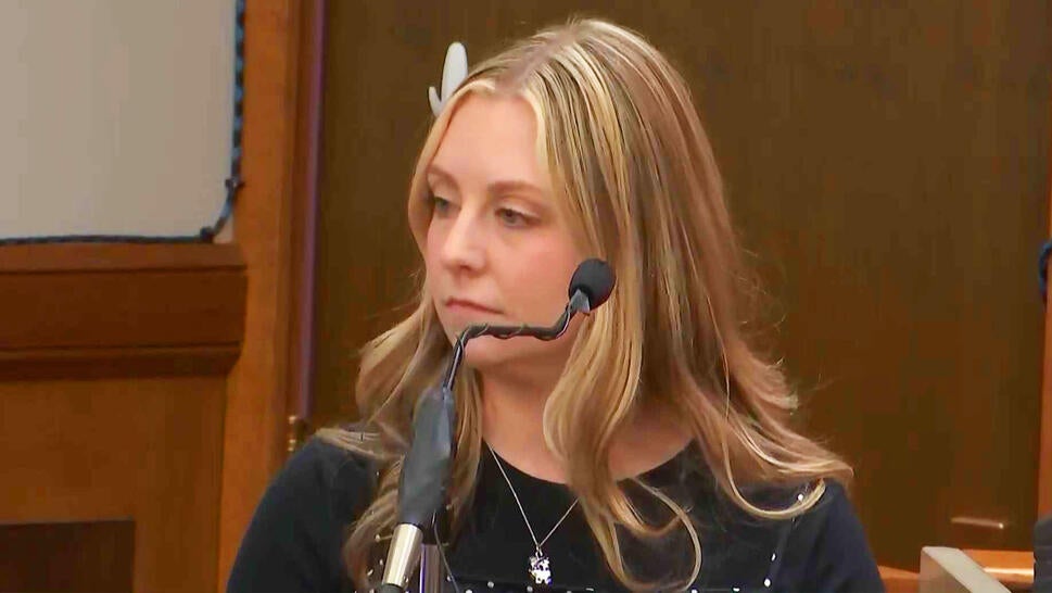 Blonde female sitting at the witness stand talking into a microphone
