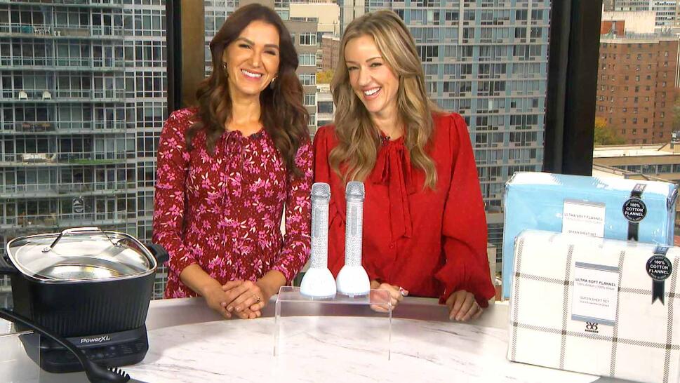 Two women standing behind a table displaying a Multi-Cooker, Karaoke Set, Sheet Set