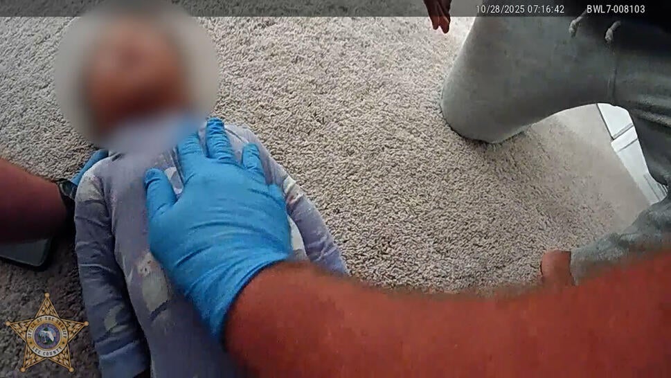 police bodycam image of infant with blurred face lying on carpeted floor, with blue-gloved hand of policeman on her chest, applying CPR