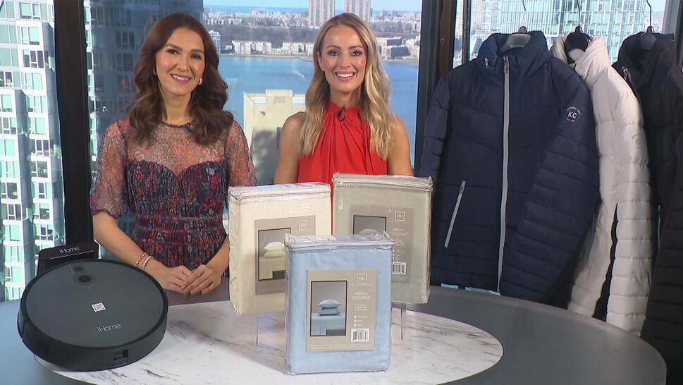 Two women standing behind a table displaying a Robot Vacuum, Sheet Set, Puffer Jacket