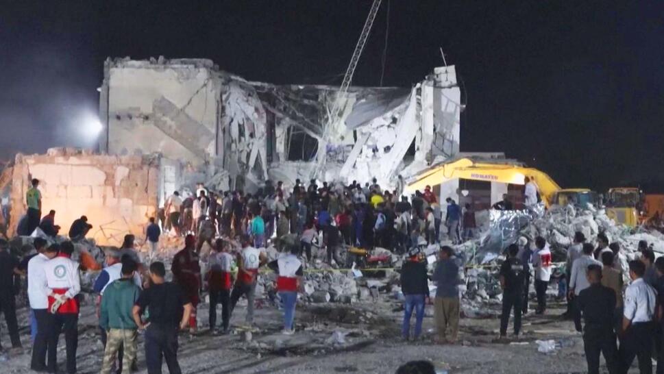 Hugely damaged cement structure surrounded by people searching through rubble