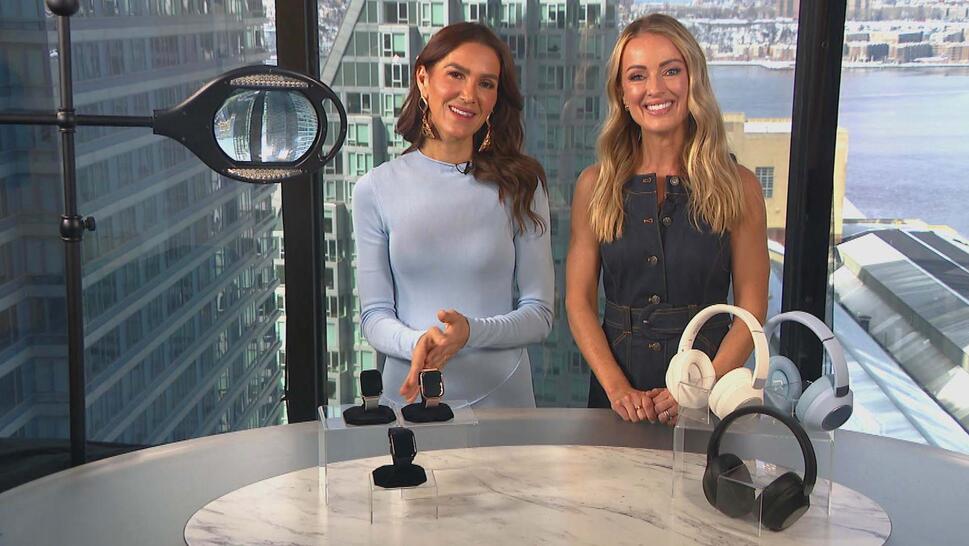Two women standing behind a table displaying a Magnifier, Smartwatch, Wireless Headphones