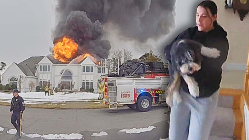 Firefighters spray water on house fire, woman carrying dog.