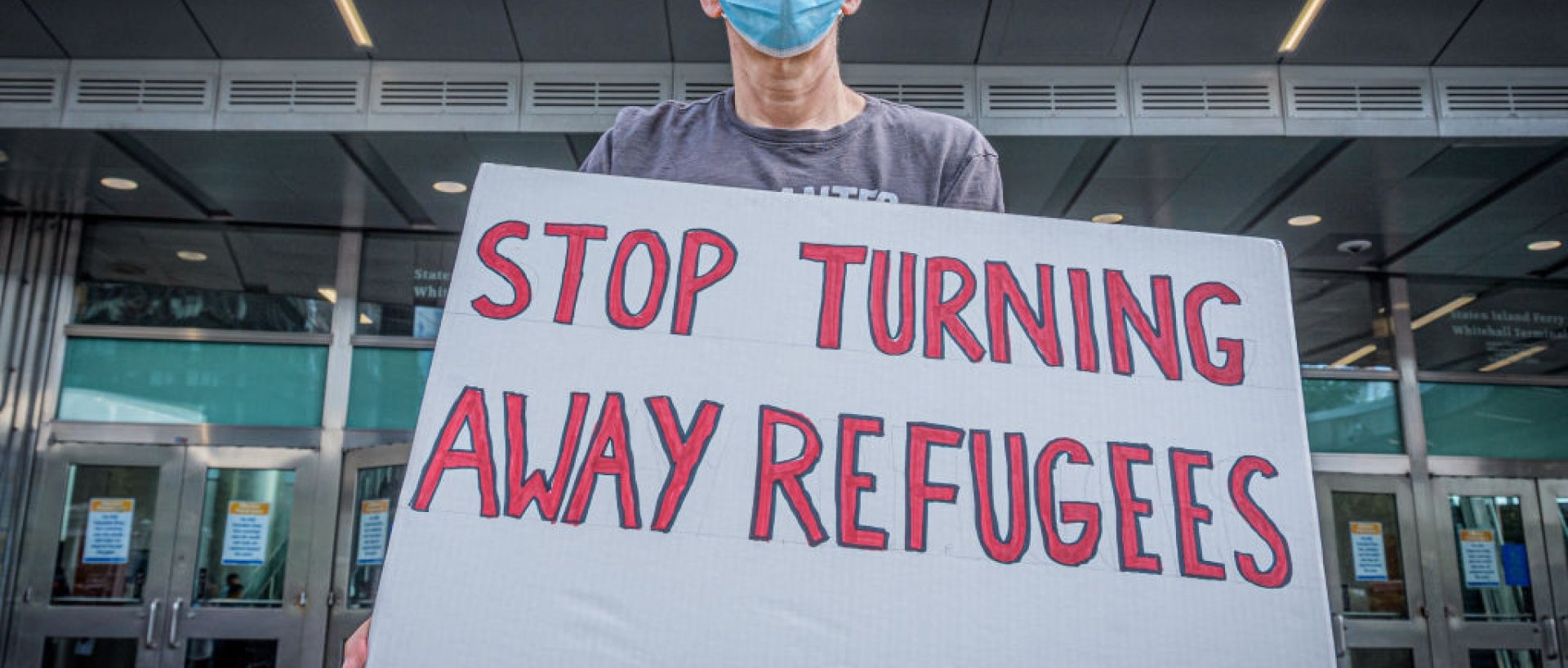 Masked man holding "stop turning away refugees" sign