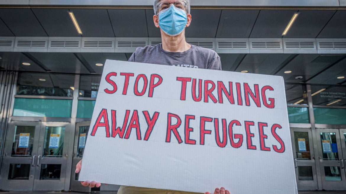 Masked man holding "stop turning away refugees" sign