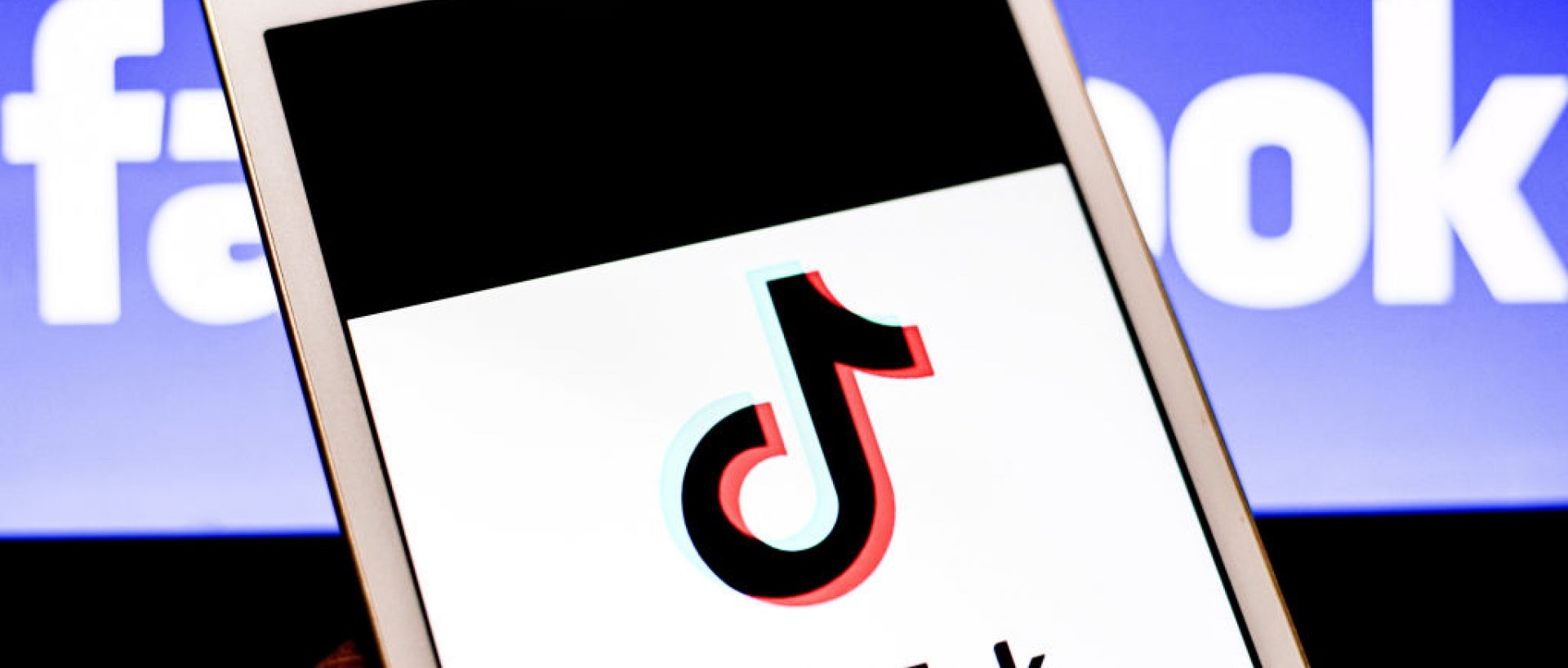 Tiktok logo on the screen of a phone being held by a hand, the facebook logo in the background