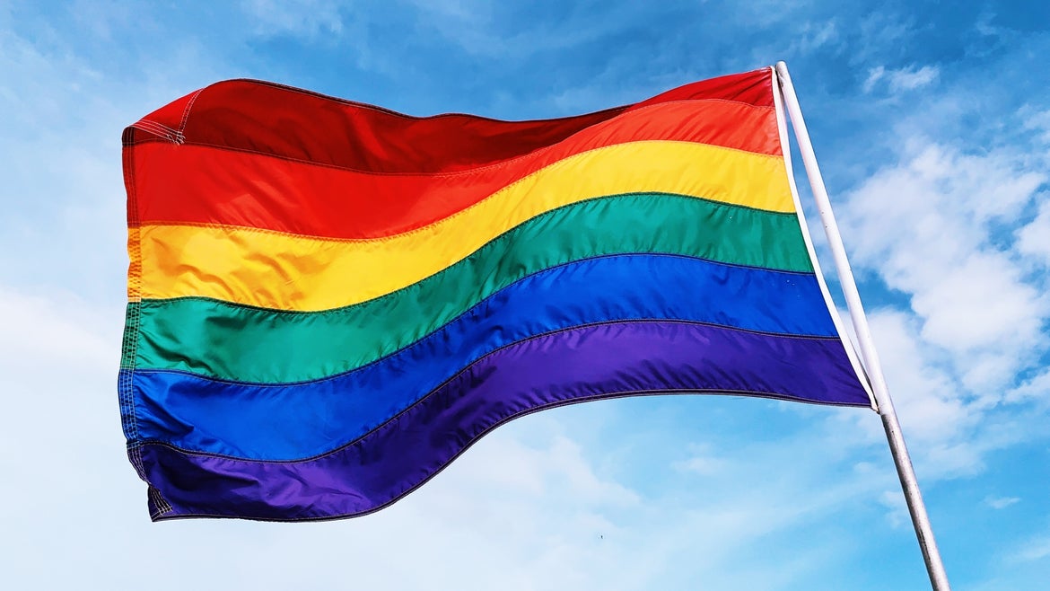 Rainbow flag signifying LGBTQ pride waving in the wind against blue sky.