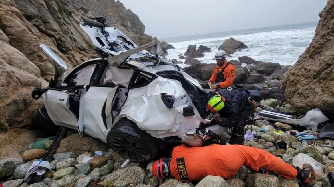 white car completely smashed at bottom on cliff, authorities in orange jumpsuits and white helmets search the car